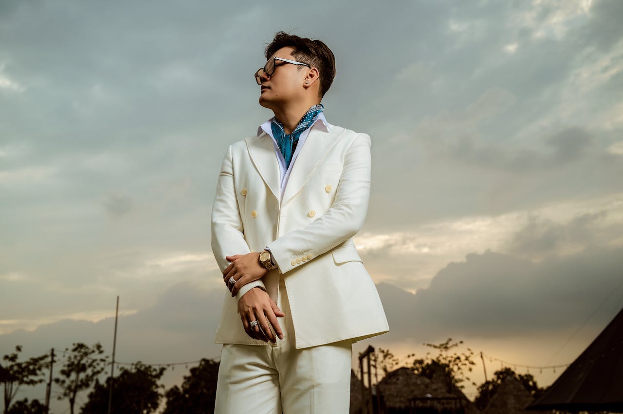Fashionable man in a white suit standing outdoors against a cloudy sky, projecting confidence and style.