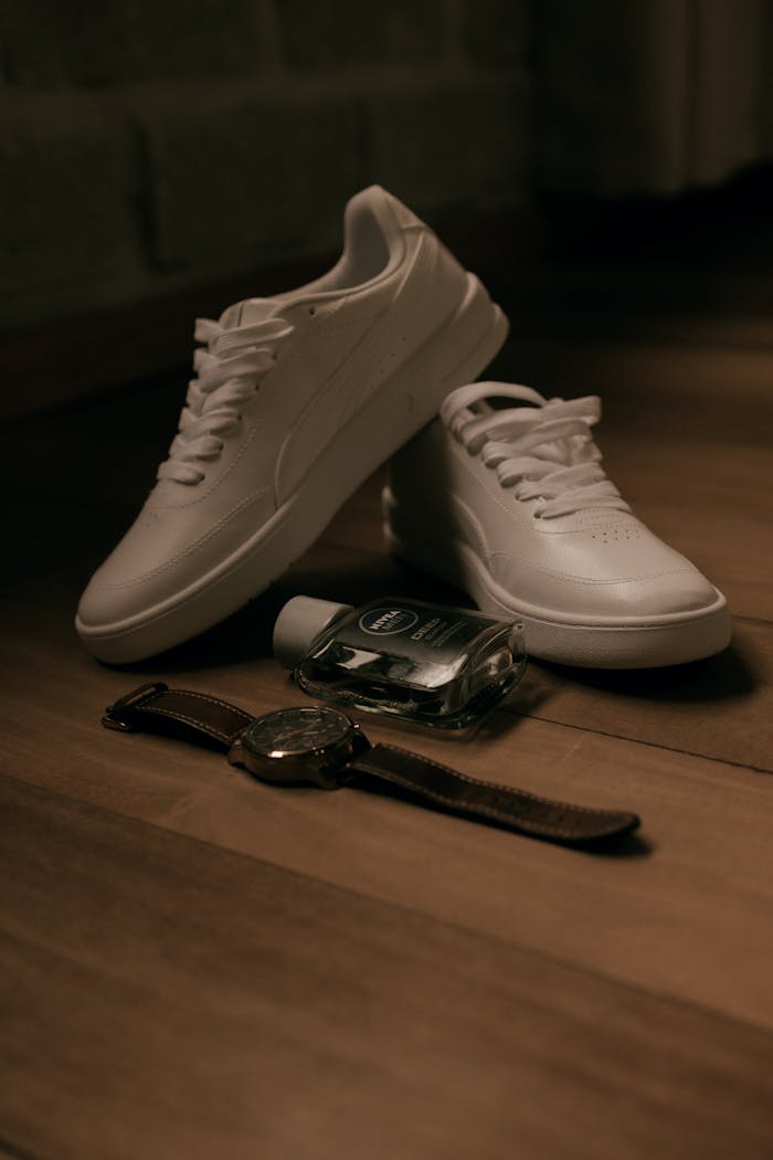 White sneakers, watch, and cologne arranged on wooden floor, warm tones.